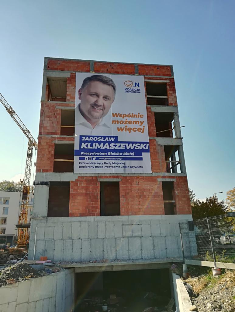 Baner wyborczy z podobizną kandydata Jarosława Klimaszewskiego umieszczony na niedokończonym budynku z czerwonej cegły, widoczny żuraw budowlany w tle.