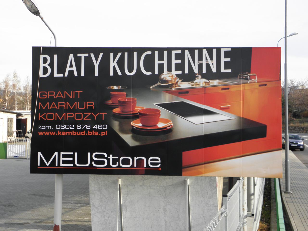 Billboard reklamowy z blatem kuchennym: granit, marmur, kompozyt. Pomarańczowe szafki, czarny blat. Napis MEUStone i dane kontaktowe.