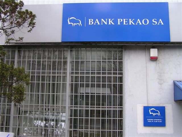 Elewacja budynku Banku Pekao SA z logo i nazwą na niebieskim szyldzie. Okna zabezpieczone kratami, mały szyld z godzinami otwarcia po prawej stronie.