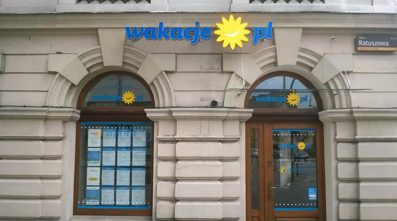 Fasada biura podróży 'wakacje.pl' z niebieskim napisem i żółtym słoneczkiem. Na oknach plakaty z ofertami. Widoczny adres: ul. Ratuszowa 5-3.
