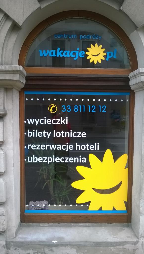 Oklejone okno biura podróży z logo 'wakacje.pl', numerem telefonu i ofertą: wycieczki, bilety lotnicze, rezerwacje hoteli, ubezpieczenia. Żółte słońce.