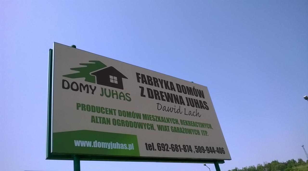 Tablica reklamowa firmy Domy Juhas z logo i danymi kontaktowymi na tle błękitnego nieba. Informacja o producentach domów z drewna, altan i wiat garażowych.