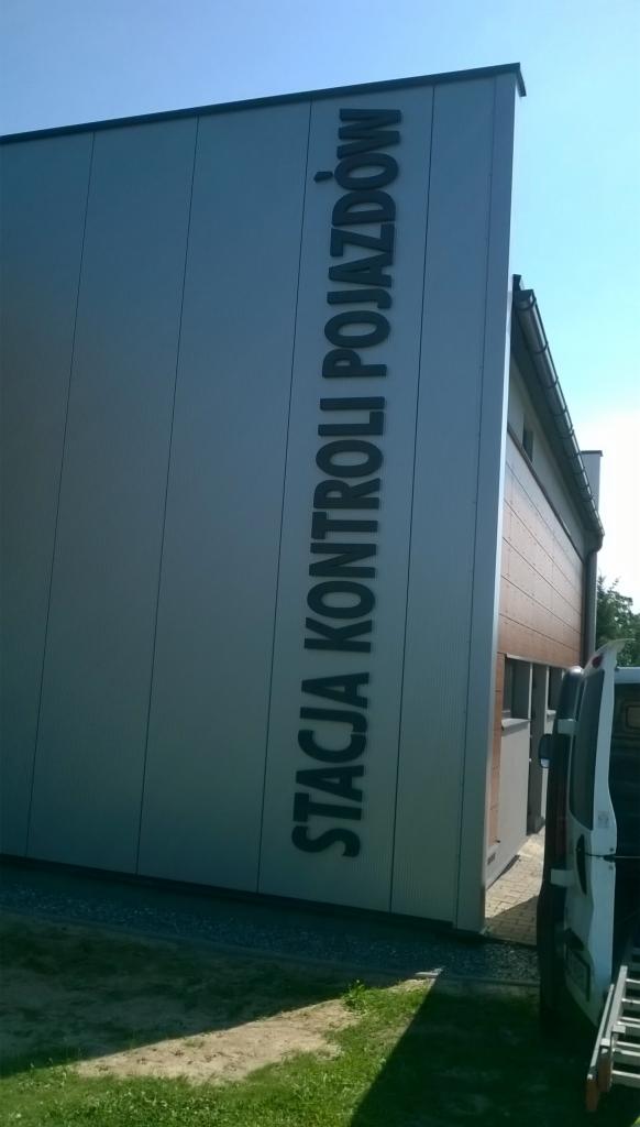 Budynek stacji kontroli pojazdów z pionowym napisem na elewacji. Obok zaparkowany biały bus. Słoneczny dzień.