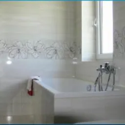 Jasna łazienka z wanną, grzejnikiem drabinkowym i dekoracyjnym pasem płytek z motywem kwiatowym. Okno z roletą w tle, minimalistyczny styl.