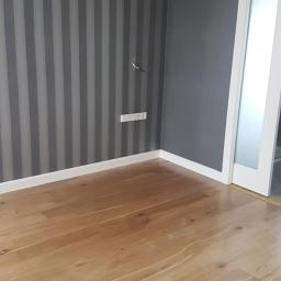 KAMP Krzysztof Bieniek - Pokój po odświeżeniu: ściana w szaro-białe pasy, gładka szara ściana, drewniana podłoga i białe listwy przypodłogowe, widoczny fragment gniazdka elektrycznego z wystającymi kablami.