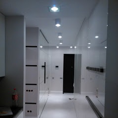 Nowoczesny przedpokój w bieli z połyskującymi panelami ściennymi, oświetleniem punktowym i minimalistyczną szafą z czarnymi akcentami.