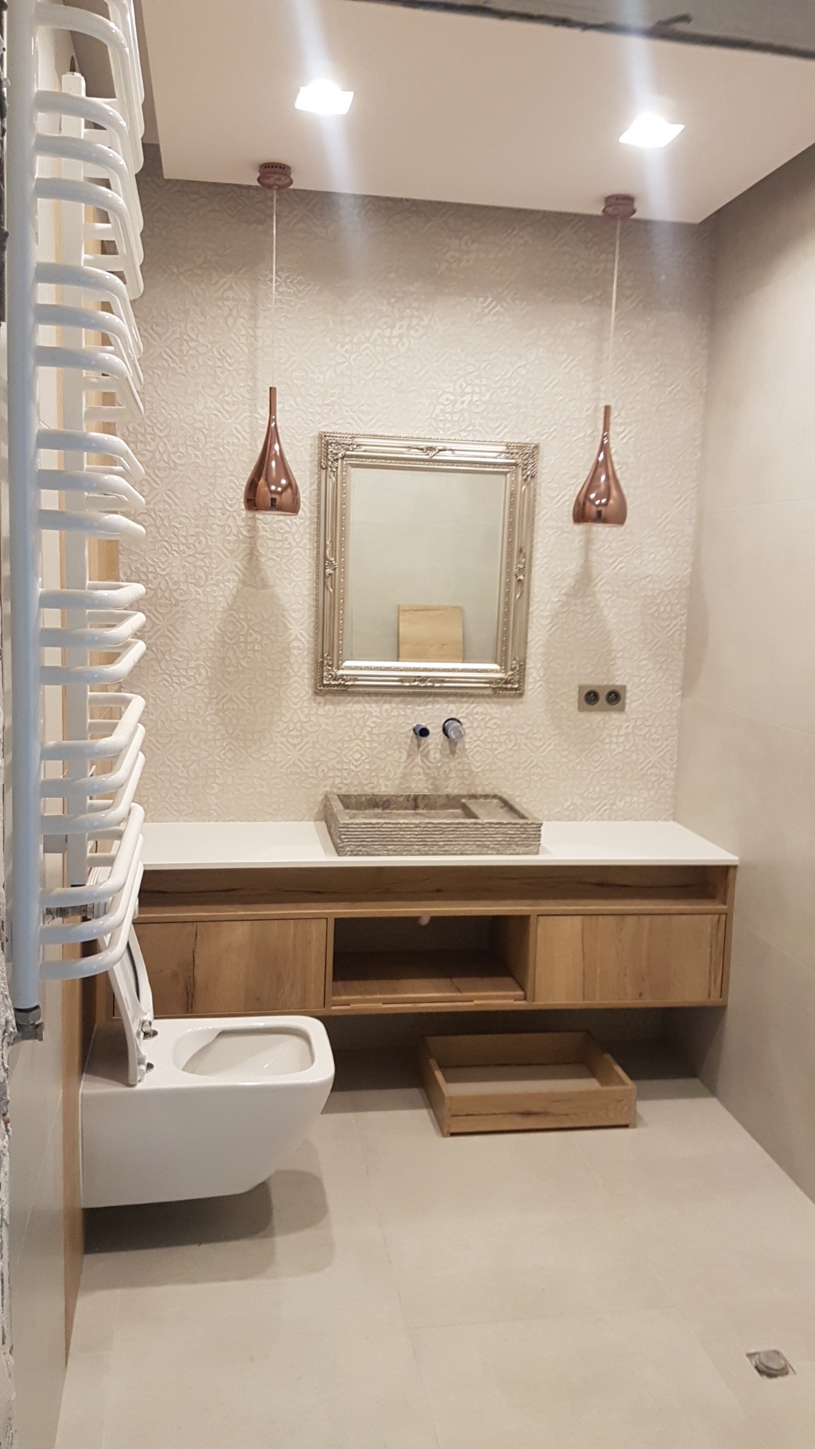 Nowoczesna łazienka z wiszącą toaletą, umywalką w dekoracyjnej ramie, drewnianą szafką pod umywalką i miedzianymi lampami wiszącymi.