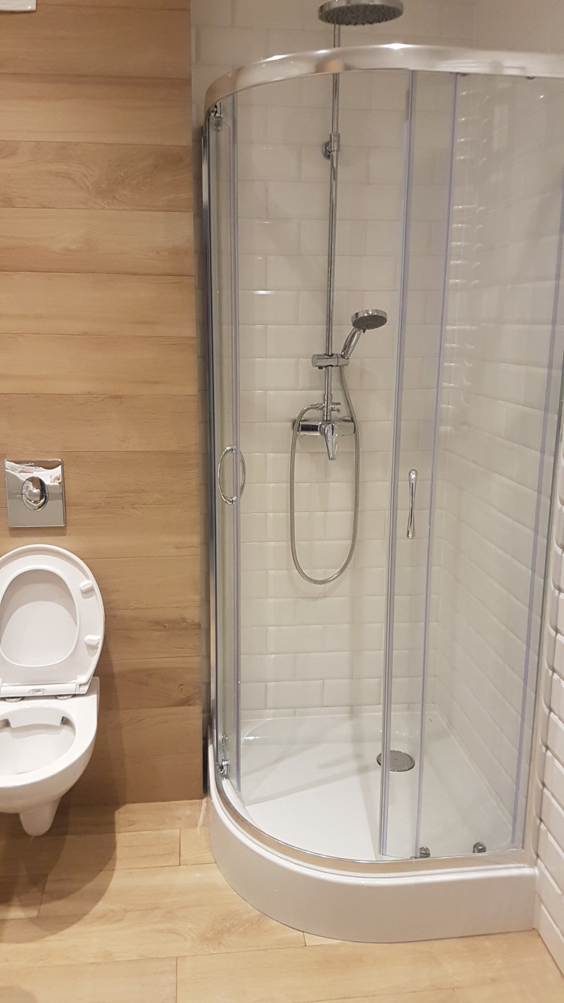 Nowoczesna łazienka z kabiną prysznicową narożną z chromowanymi detalami, wiszącą toaletą z otwartą klapą i drewnopodobnymi płytkami na ścianach i podłodze.