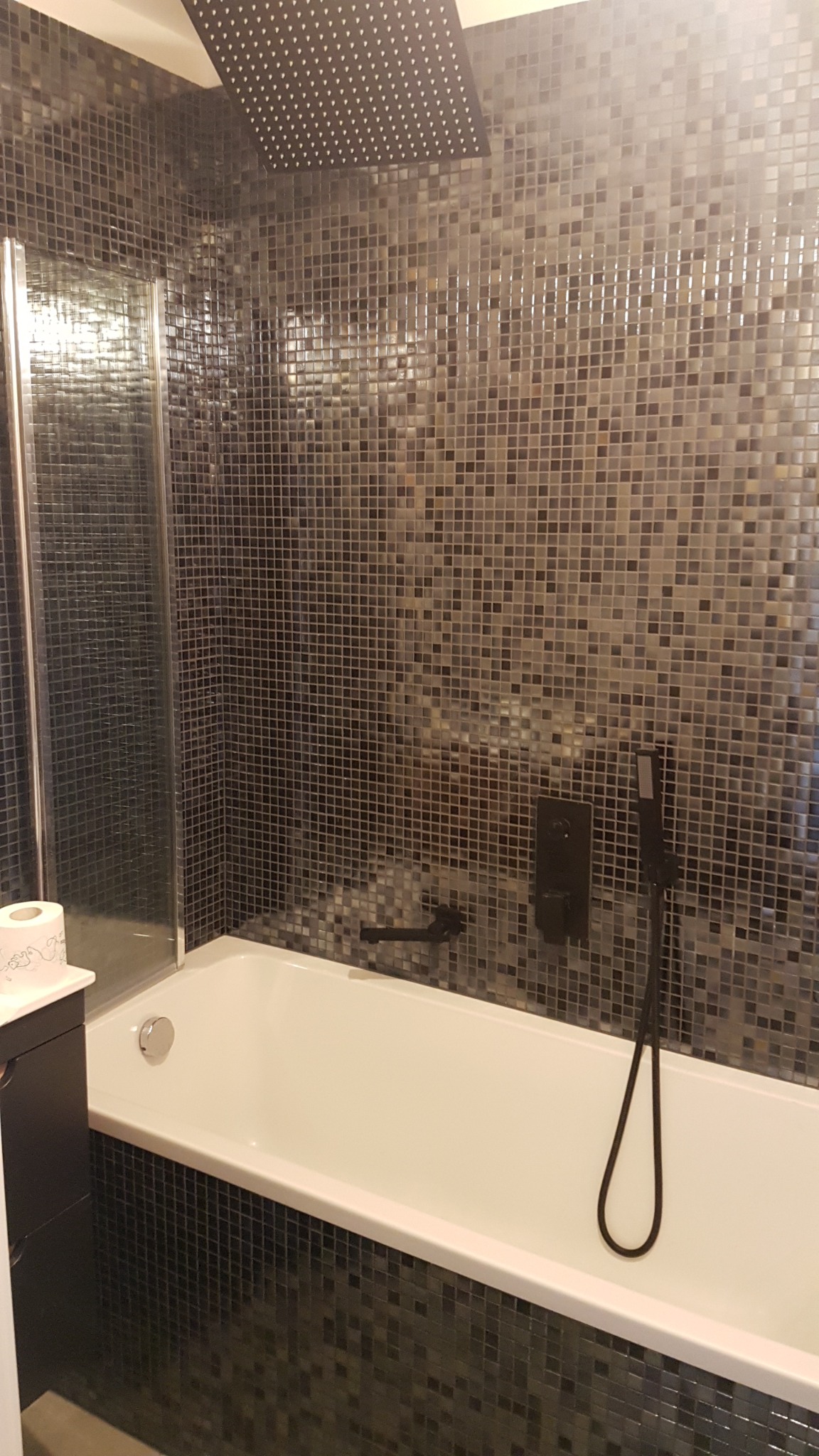 Biała wanna z czarną armaturą i deszczownicą na tle ściany wyłożonej mozaiką w odcieniach szarości i czerni, kabina prysznicowa po lewej stronie.