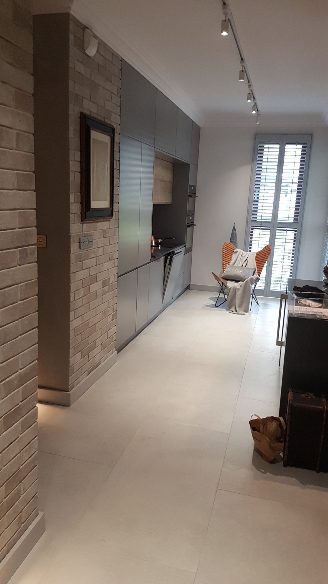 Nowoczesna kuchnia z szarymi szafkami, ceglaną ścianą i jasną podłogą. Widoczny fragment jadalni z designerskim krzesłem i dekoracyjnymi przedmiotami.