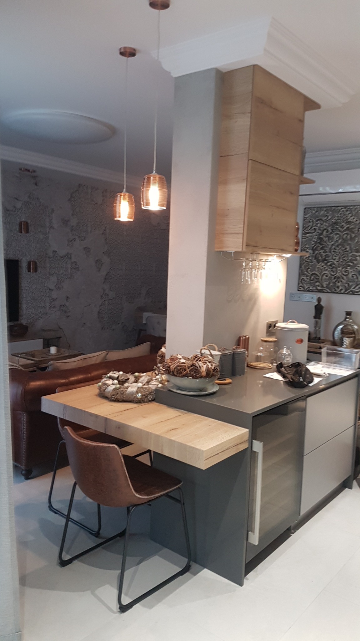 Częściowo widoczna kuchnia z blatem barowym z jasnego drewna, brązowe krzesło barowe, dekoracje świąteczne na blacie, w tle salon z brązową skórzaną sofą i szarą ścianą z fakturą.