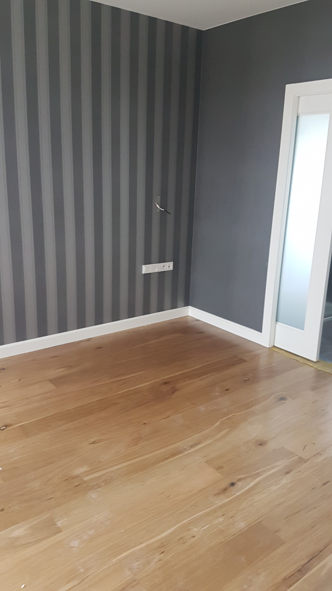 Pokój po odświeżeniu: ściana w szaro-białe pasy, gładka szara ściana, drewniana podłoga i białe listwy przypodłogowe, widoczny fragment gniazdka elektrycznego z wystającymi kablami.