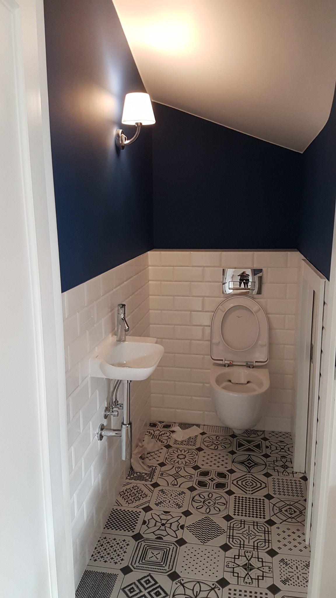 Mała, odnowiona łazienka w Warszawie z białą umywalką, toaletą i wzorzystą podłogą w stylu patchwork, oświetlona kinkietem na granatowej ścianie.
