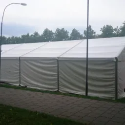 Biały namiot imprezowy z widocznymi lampami ulicznymi i budynkami w tle, ustawiony na brukowanym chodniku obok zieleni miejskiej.