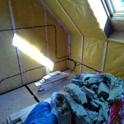 Domy Baca - Poddasze w trakcie prac wykończeniowych: widoczne ocieplenie folią i wełną mineralną, instalacja elektryczna, okno dachowe oraz przygotowane deski podłogowe. Częściowe oświetlenie słoneczne przez...