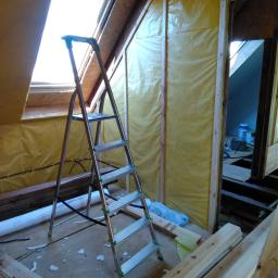 Domy Baca - Wnętrze poddasza w trakcie ocieplania, widoczna drabina aluminiowa, okno dachowe, żółta folia izolacyjna na ścianach, przewody elektryczne oraz fragment podłogi z resztkami materiałów budowlanych.