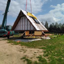 Domy Baca - W Obrytem, dźwig podnosi prefabrykowany dach z oknem dachowym, pokryty membraną i łatami, w tle widoczny szkielet domu.