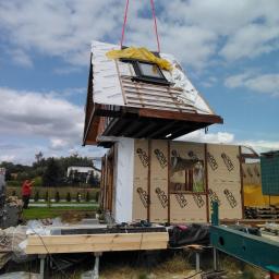 Domy Baca - Podnoszenie dachu domu szkieletowego za pomocą dźwigu, widoczne elementy konstrukcyjne i folia ochronna na dachu, w tle budynek mieszkalny.