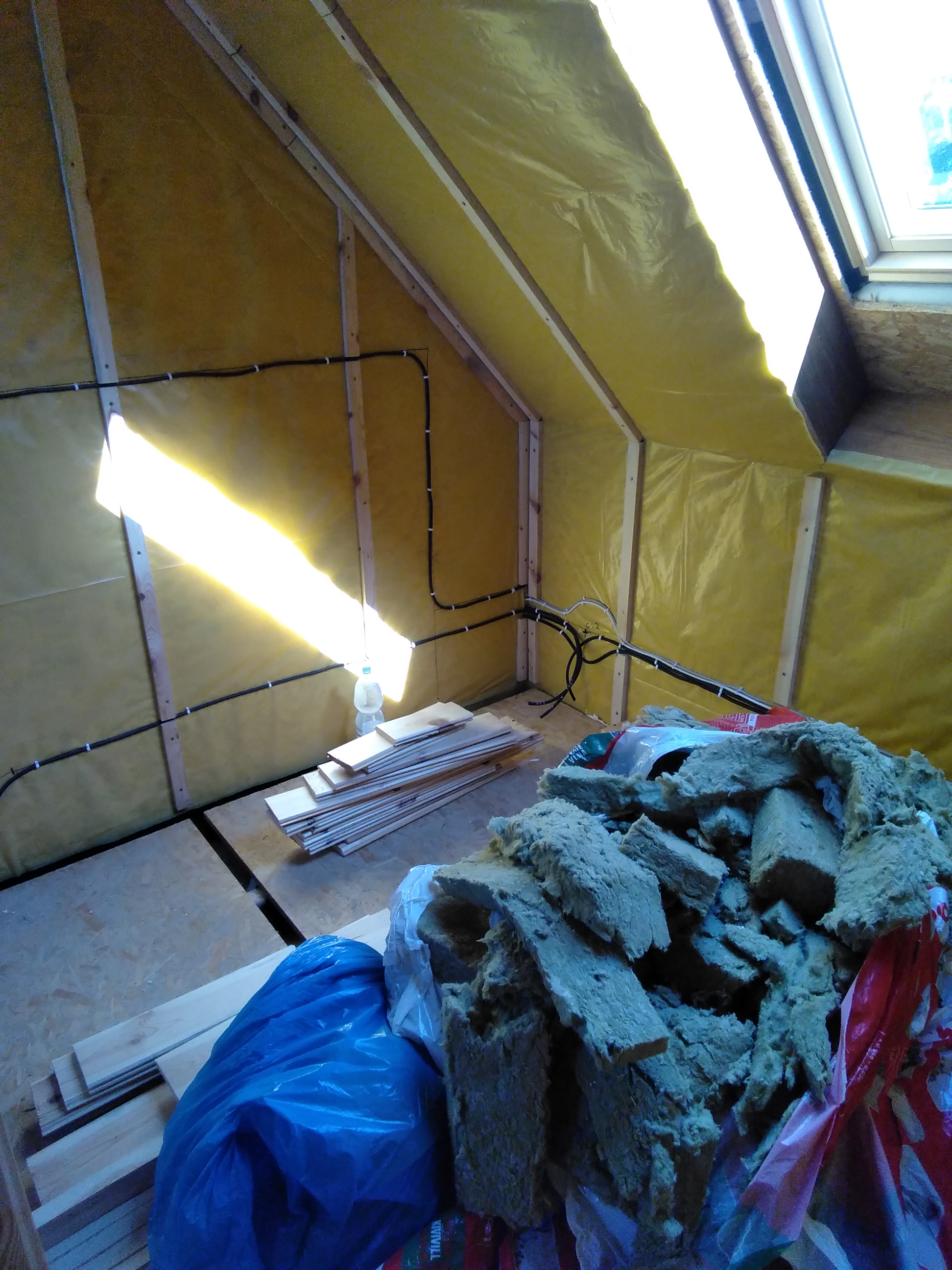 Poddasze w trakcie prac wykończeniowych: widoczne ocieplenie folią i wełną mineralną, instalacja elektryczna, okno dachowe oraz przygotowane deski podłogowe. Częściowe oświetlenie słoneczne przez...