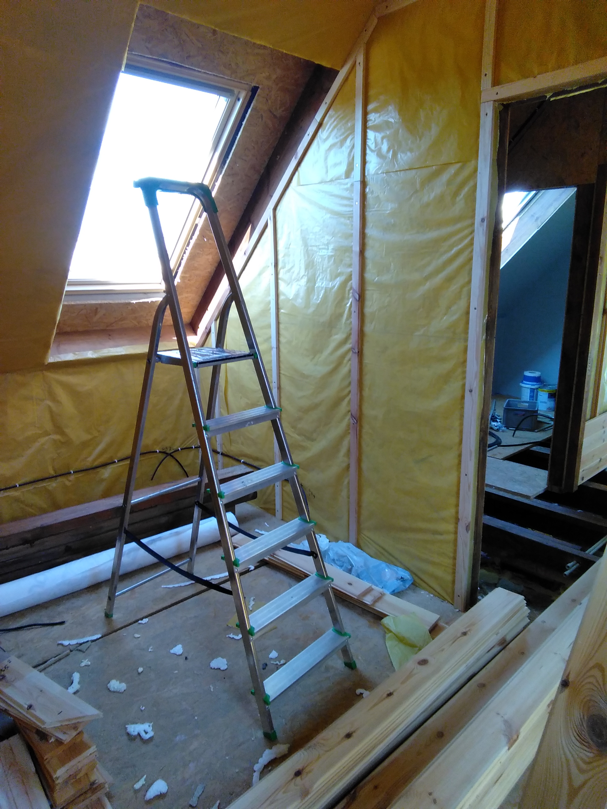 Wnętrze poddasza w trakcie ocieplania, widoczna drabina aluminiowa, okno dachowe, żółta folia izolacyjna na ścianach, przewody elektryczne oraz fragment podłogi z resztkami materiałów budowlanych.