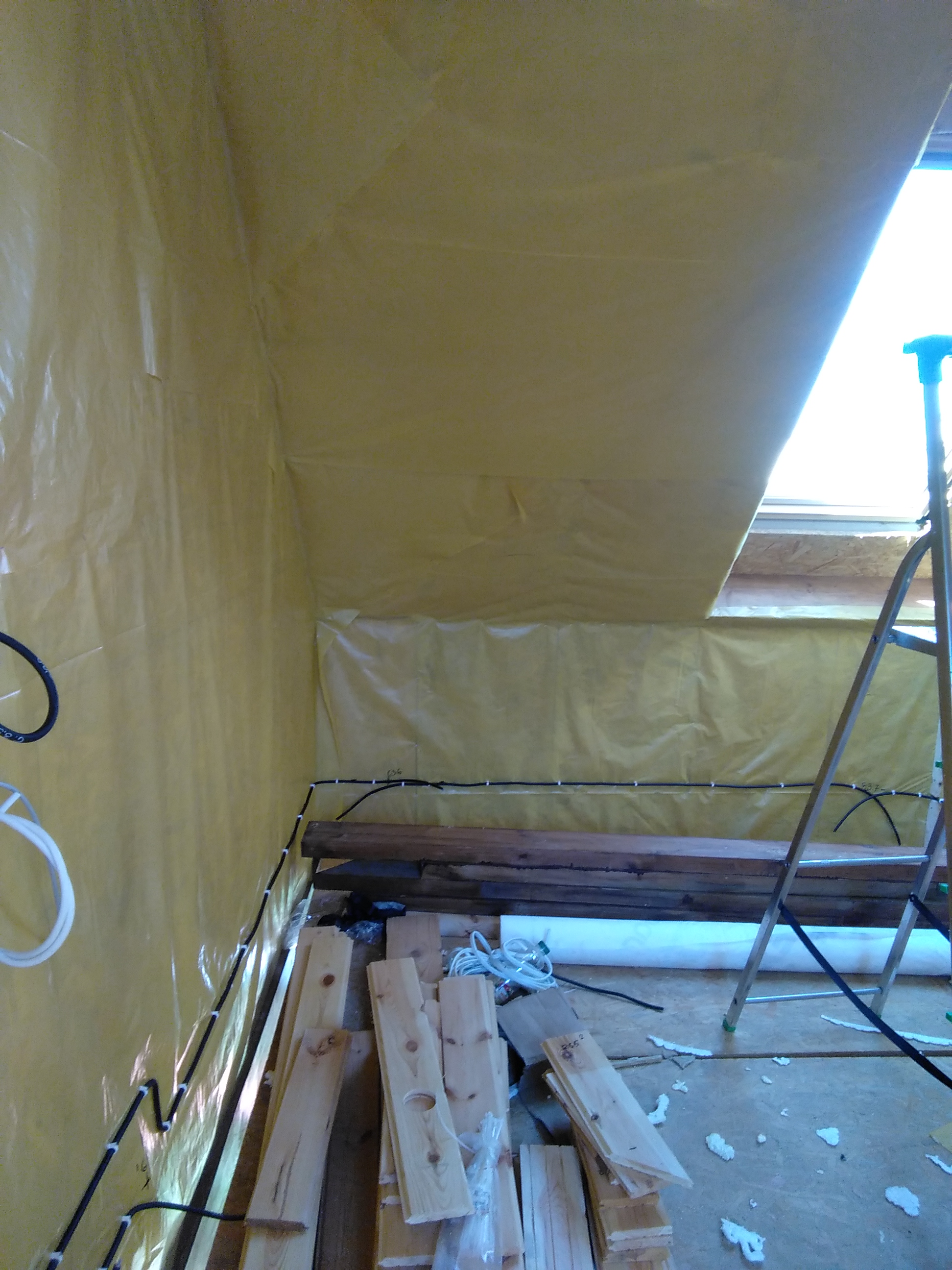 Wnętrze domu szkieletowego w trakcie montażu dachu z izolacją folią paroizolacyjną, widoczne instalacje elektryczne prowadzone po ścianach i drewniane elementy konstrukcyjne oraz drabina.