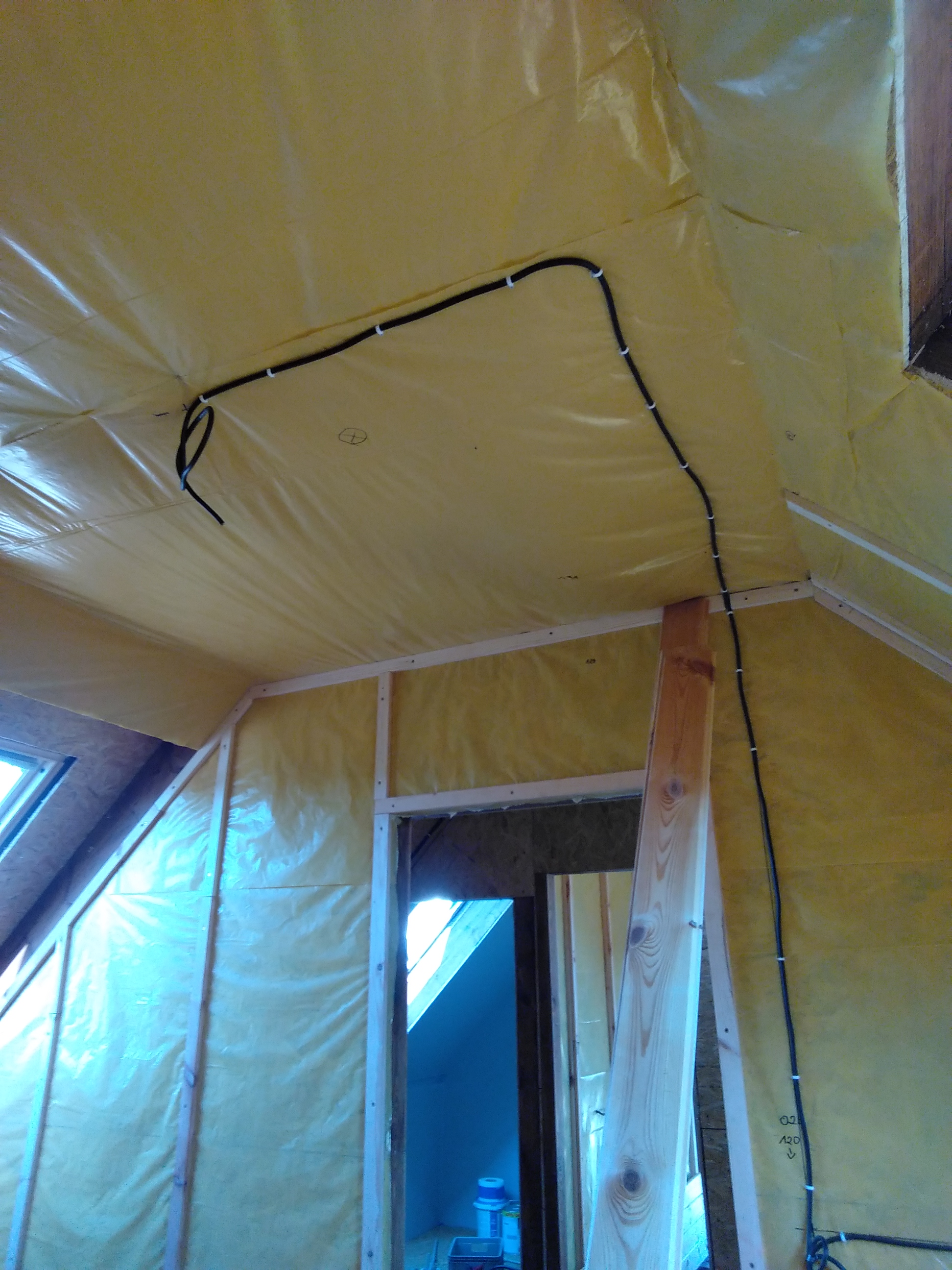 Wnętrze poddasza w trakcie budowy, widoczne drewniane elementy konstrukcyjne, żółta folia izolacyjna oraz poprowadzona instalacja elektryczna z użyciem białych uchwytów kablowych.