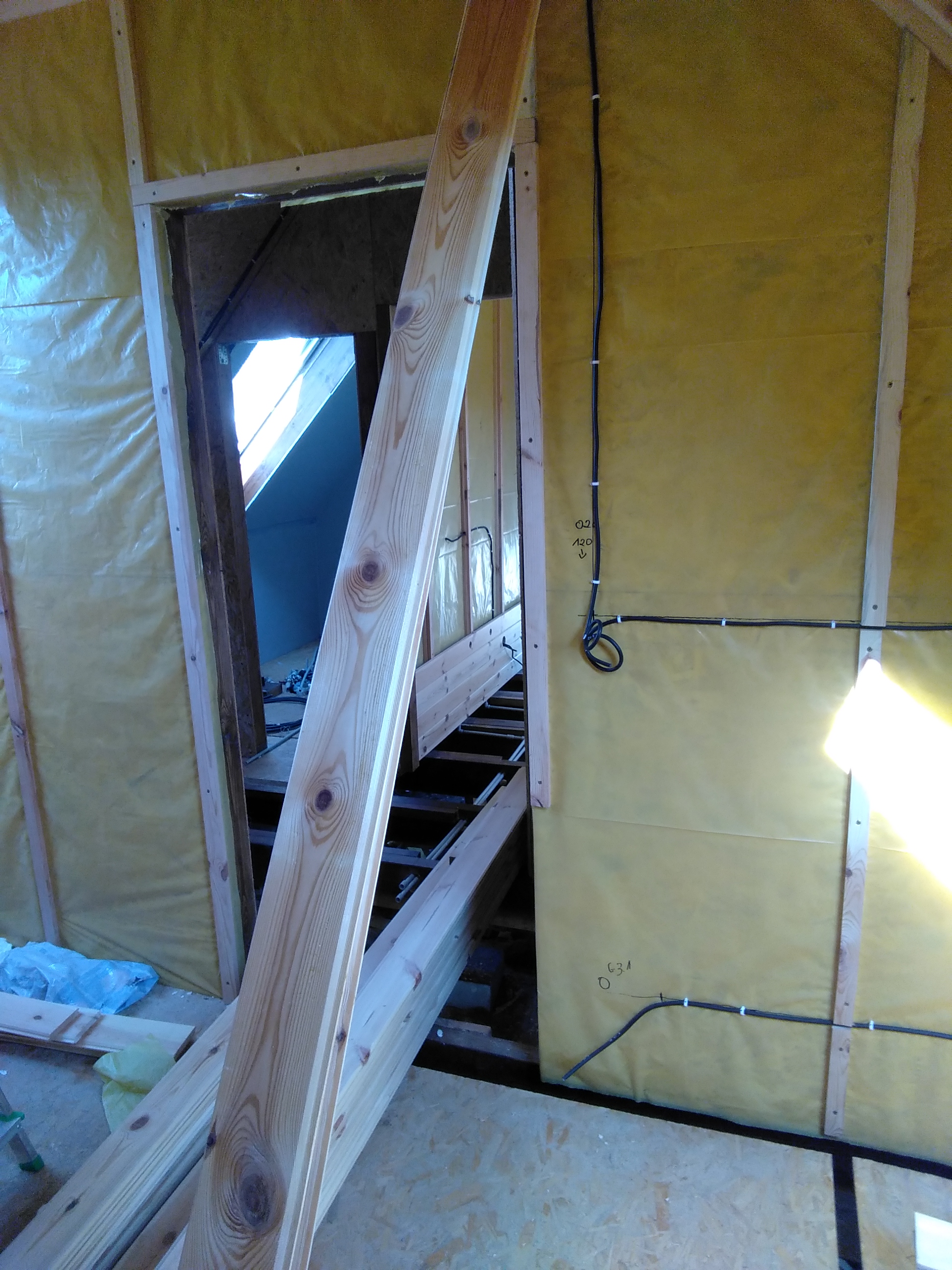 Wnętrze poddasza w trakcie budowy, widoczne drewniane elementy konstrukcyjne, izolacja folią, instalacja elektryczna prowadzona po ścianie i fragment okna dachowego.