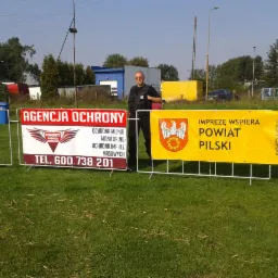 Mężczyzna w kamizelce ochroniarskiej stoi między dwoma banerami: Agencja Ochrony Combat i Powiat Pilski wspierający imprezę. Widok na zewnątrz, trawa.