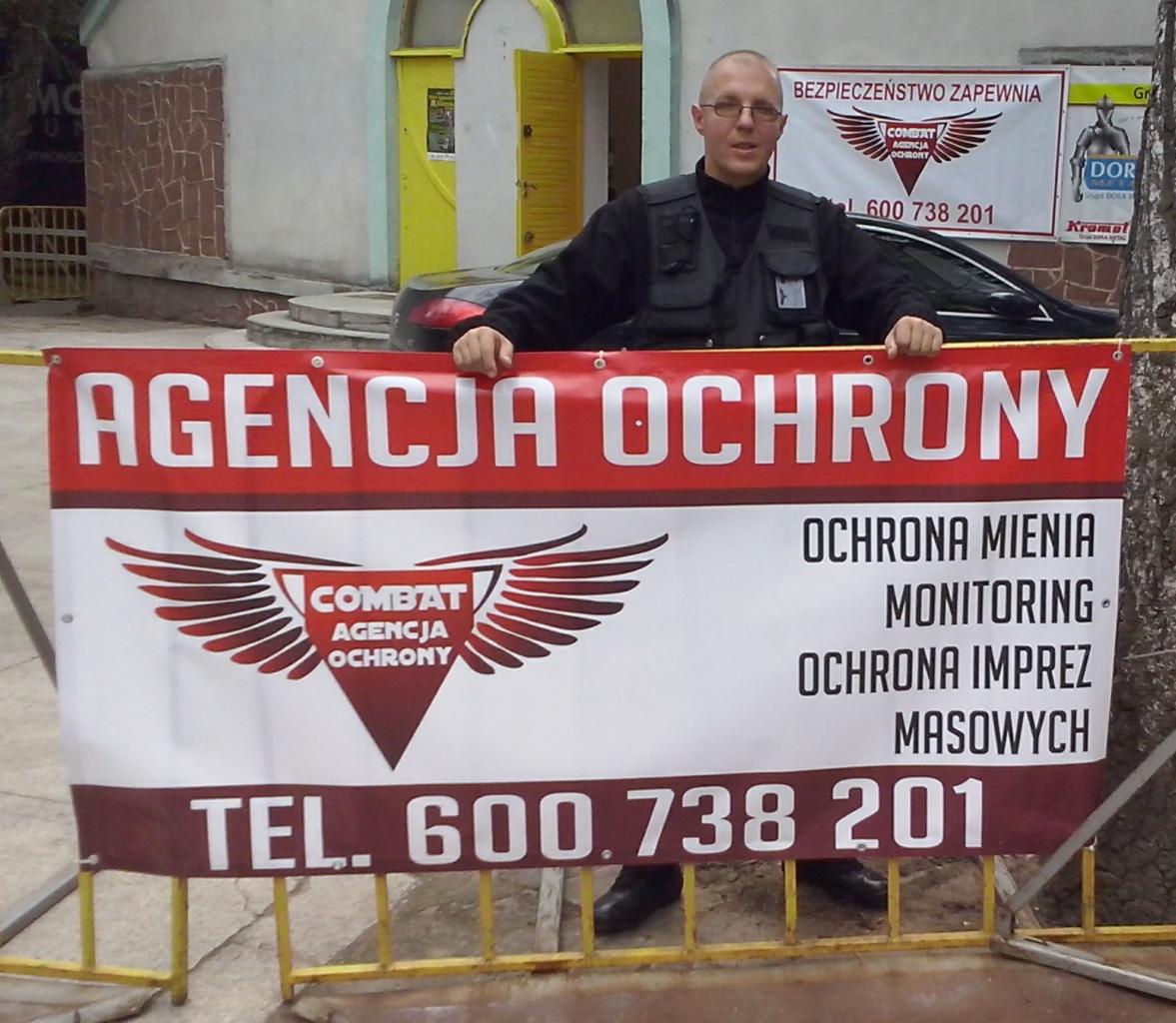Mężczyzna w kamizelce ochronnej prezentuje baner agencji ochrony Combat z ofertą: ochrona mienia, monitoring, ochrona imprez masowych. W tle budynek i zaparkowany samochód.
