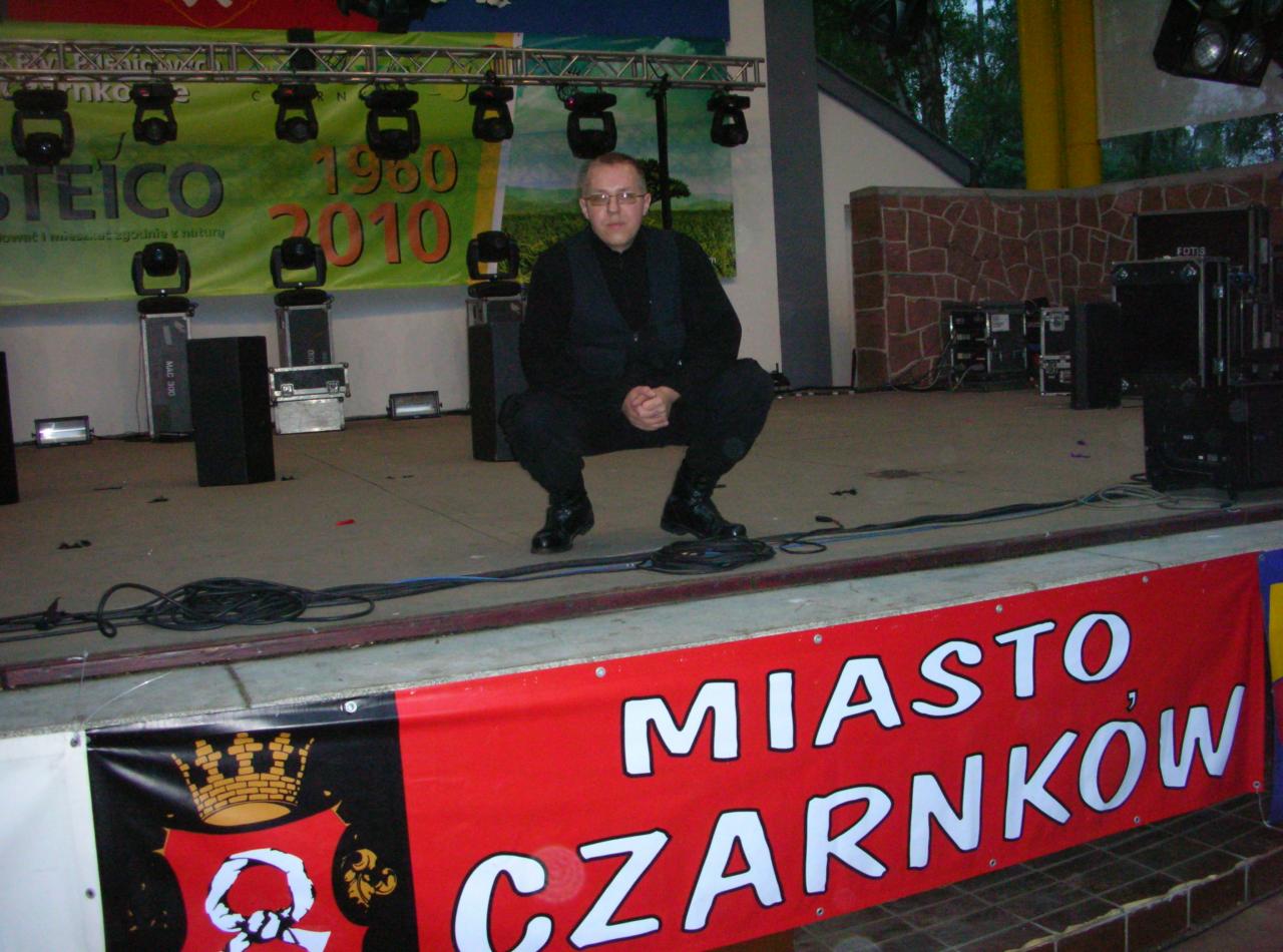 Mężczyzna w czarnym stroju kuca na scenie przed banerem 'Miasto Czarnków', otoczony sprzętem nagłośnieniowym i oświetleniowym.