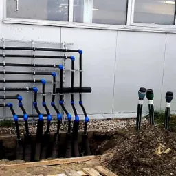 Czarny system rur z niebieskimi złączkami przymocowany do ściany budynku, część rur wchodzi do wykopu w ziemi, a pozostałe wystają z ziemi z zaślepkami.