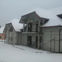 Dom w trakcie elewacji zimą, pokryty śniegiem, z widocznymi rusztowaniami, w szary dzień. Częściowo otynkowany, z elementami wykończenia dachu.