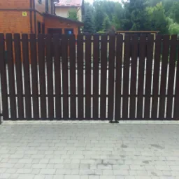 Ciemnobrązowe, drewnopodobne ogrodzenie posesji w Aleksandrowie Łódzkim z furtką i skrzynką na listy, widok z zewnątrz na kostkę brukową.