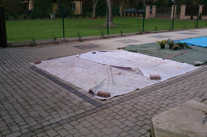 Przygotowanie podłoża pod budowę: biała plandeka przyciśnięta cegłami na kostce brukowej, obok zielona plandeka z roślinami w doniczkach, w tle trawnik i ogrodzenie z siatki.