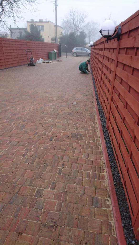 Czerwona kostka brukowa na podjeździe, nowy drewniany płot z kulistymi lampami, pracownik w zielonym kombinezonie przy płocie, widoczne narzędzia i worki w tle.