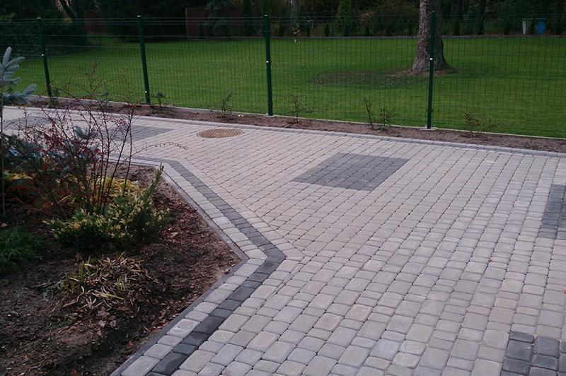 Ułożona kostka brukowa w odcieniach szarości z ciemniejszym obramowaniem, widoczny fragment ogrodu z zielonym trawnikiem i metalowe ogrodzenie panelowe.