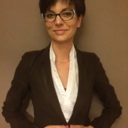 Uśmiechnięta kobieta w okularach i brązowej marynarce na tle beżowej ściany. Ręce złożone na wysokości brzucha, widoczny zegarek na nadgarstku.