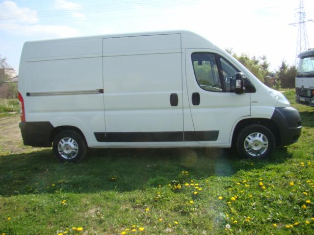 Biały furgon dostawczy Fiat Ducato z przesuwanymi drzwiami, zaparkowany na trawniku z żółtymi kwiatami, w tle słup wysokiego napięcia i fragment innego pojazdu.