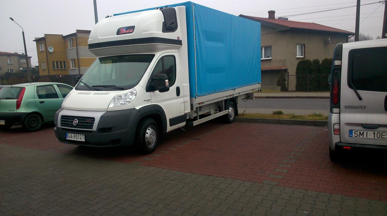 Biały samochód dostawczy Fiat Ducato z niebieską plandeką na parkingu z kostki, w tle budynki mieszkalne i zaparkowane auta.