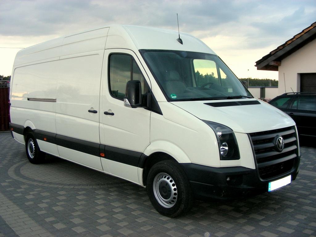 Biały bus Volkswagen Crafter na kostce brukowej, widok z boku, zarys budynku w tle. Pojazd gotowy do transportu, czysty i zadbany.