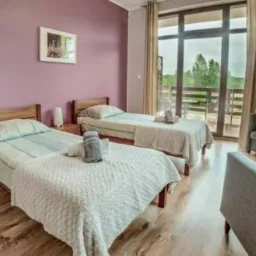 Jasny pokój z dwoma łóżkami pojedynczymi, balkonem i widokiem na zieleń, Brodnica Dolna. Ręczniki ułożone na łóżkach, telewizor na ścianie.