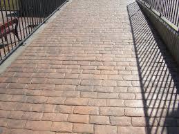 Ułożona kostka brukowa na podjeździe, widok z góry. Cień barierki na nawierzchni. W oddali widoczna ławka i fragment ogrodzenia. Jasne, słoneczne oświetlenie.