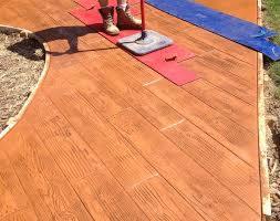 Stemplowany beton imitujący drewnianą podłogę ułożoną w jodełkę. Osoba w butach roboczych z narzędziem do stemplowania. Czerwona i niebieska folia zabezpieczająca.