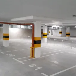 Nowoczesny parking podziemny z szarą posadzką, białymi liniami oznaczającymi miejsca postojowe i słupami pomalowanymi w czarno-żółte pasy. Widoczne rury wentylacyjne pod sufitem i numeracja miejsc...