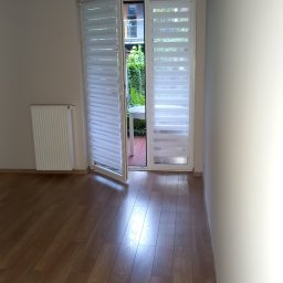 Zygmunt Gnat - Jasna sypialnia po remoncie: drewniana podłoga, białe ściany, grzejnik i drzwi balkonowe z roletami w kasecie, otwarte na zewnątrz z widokiem na zieleń.
