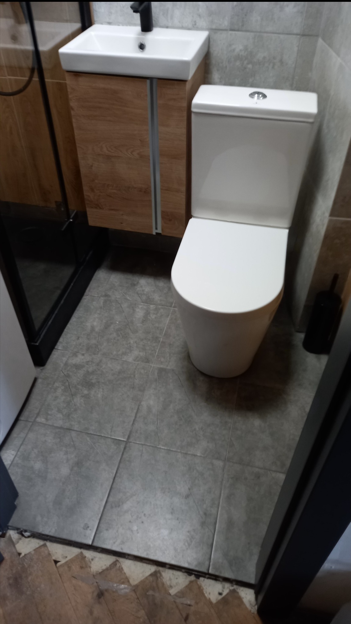 Nowoczesna toaleta z podwieszaną szafką umywalkową z drewnopodobnym frontem i czarną armaturą, szare płytki podłogowe i ścienne, widoczne fragmenty drewnianej podłogi przy progu.