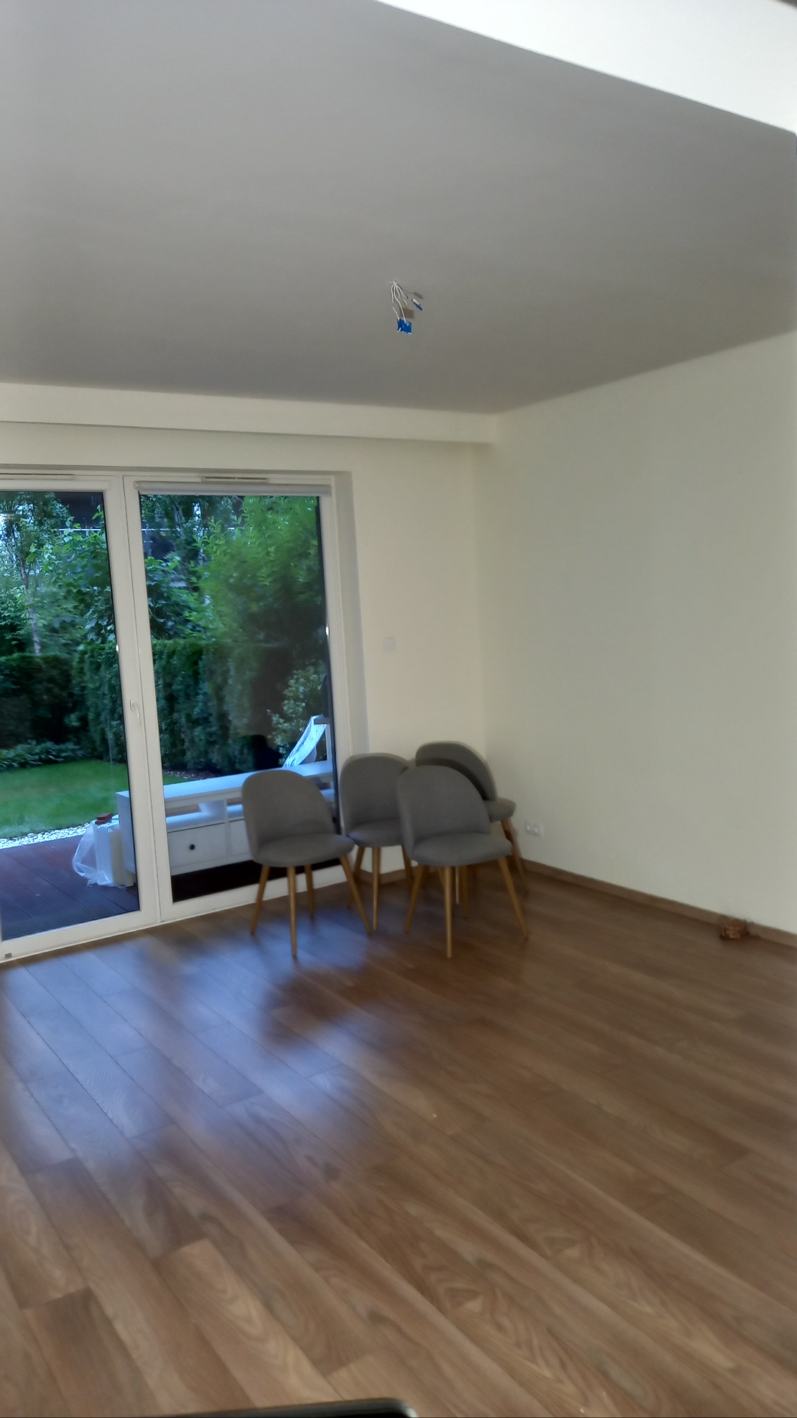 Jasny salon z drewnianą podłogą i czterema szarymi krzesłami. Widoczne przewody elektryczne wystające z sufitu, duże okno z widokiem na ogród.