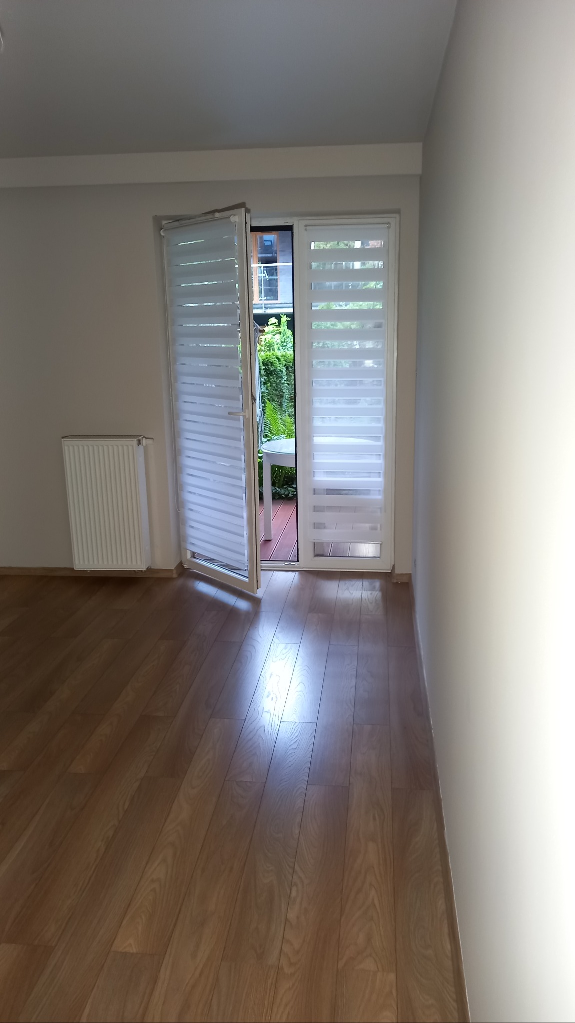 Jasna sypialnia po remoncie: drewniana podłoga, białe ściany, grzejnik i drzwi balkonowe z roletami w kasecie, otwarte na zewnątrz z widokiem na zieleń.