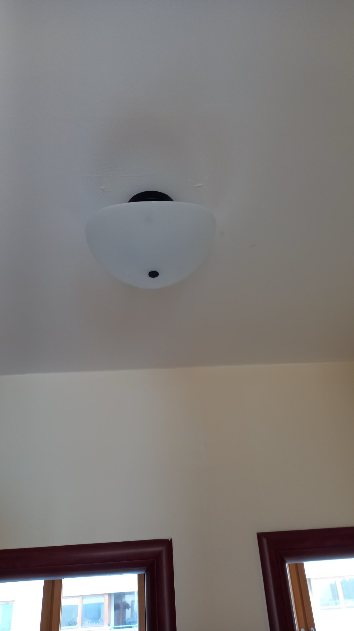 Zamontowana lampa sufitowa z mlecznego szkła, widoczna na tle pomalowanego sufitu. Częściowo widoczne okna w tle. Widok z dołu.