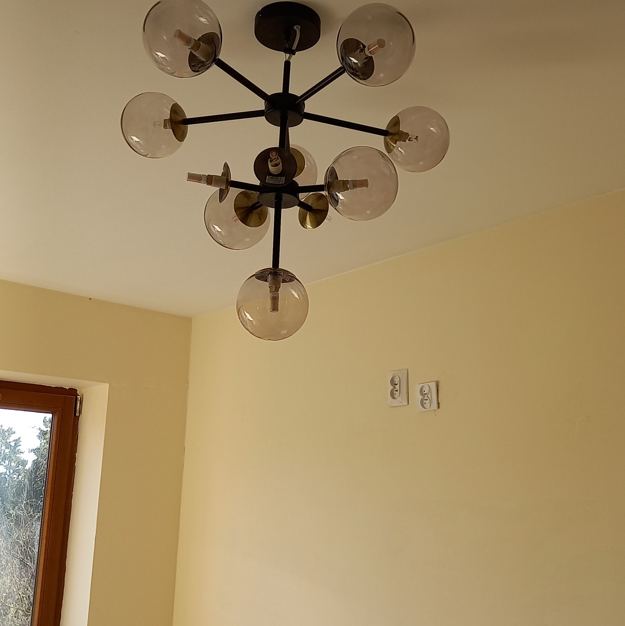 Nowoczesny żyrandol z kulistymi kloszami, zamontowany na suficie w pokoju o kremowych ścianach. Widoczne gniazdka elektryczne i okno z widokiem na drzewa.